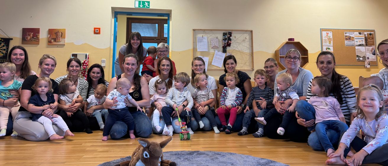 Eine Gruppe von Erwachsenen und Kindern in einem Raum mit Holzboden, einige sitzen auf dem Boden und andere stehen, bunte Flaggen hängen von der Decke und eine Pinnwand an der Wand.