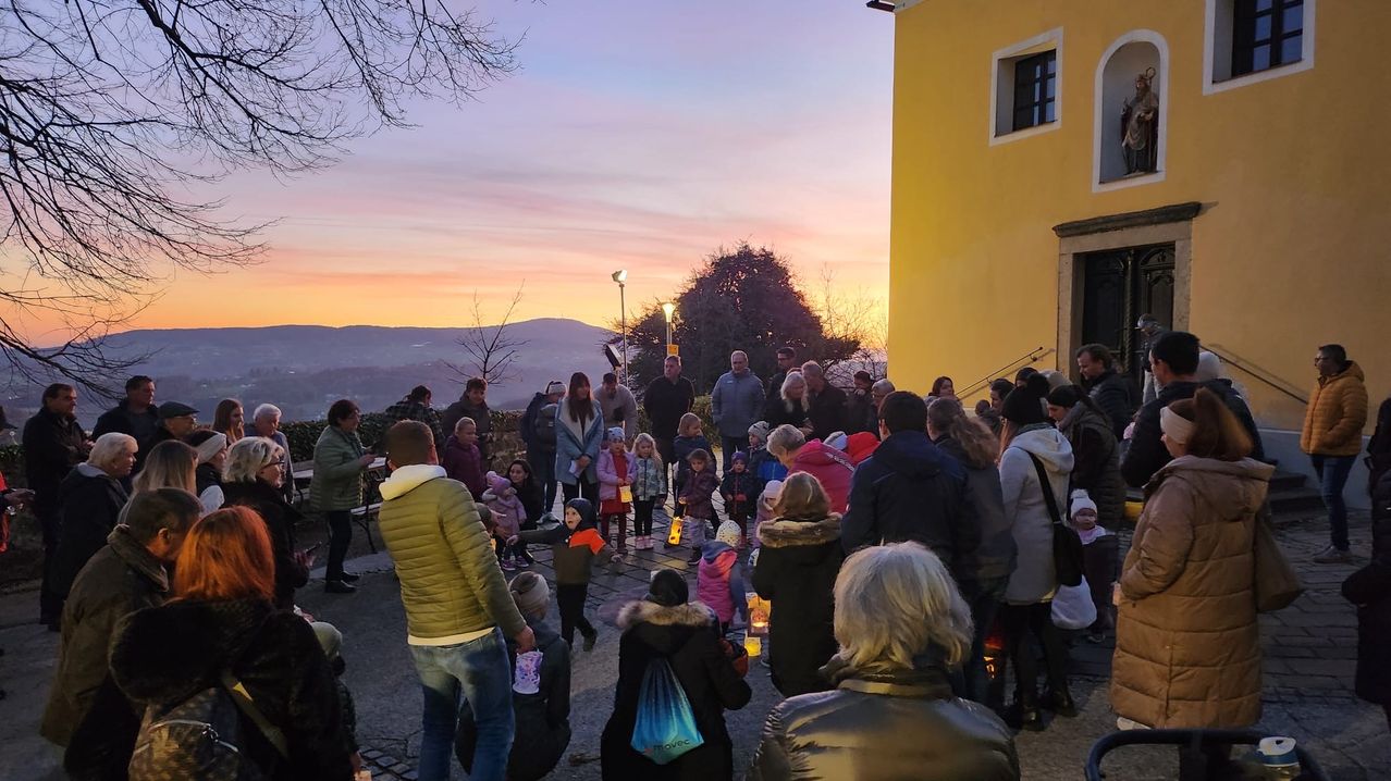 Bei Sonnenuntergang versammeln sich eine Menschenmenge, darunter Kinder mit Kerzen, vor einem gelben Gebäude. Der Himmel ist orange und pink, und es gibt Bäume und Berge in der Ferne.