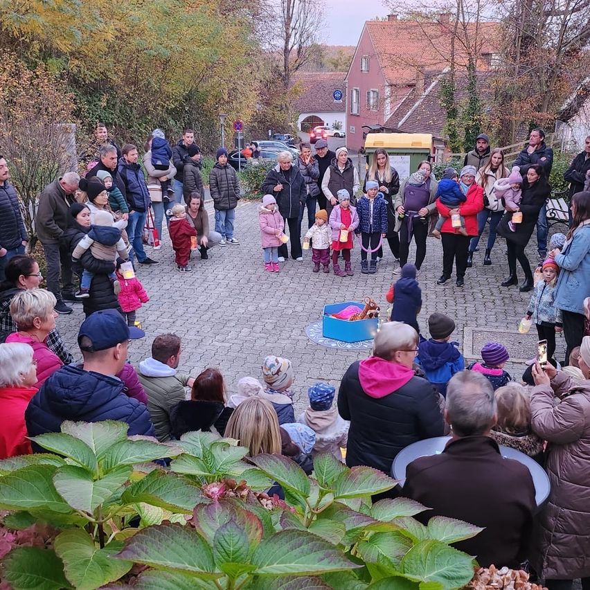 Eine Gemeindeversammlung mit Familien und Kindern findet in einem Außenbereich statt. Sie stehen im Kreis und manche halten Laternen. Im Hintergrund sind Gebäude und Autos zu sehen.