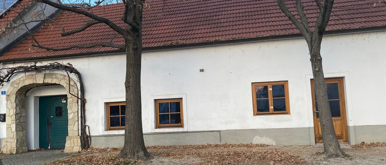 Ein altes weißes Gebäude mit rotem Dach und drei Fenstern, von denen zwei offen sind. Vor dem Gebäude stehen zwei Bäume.