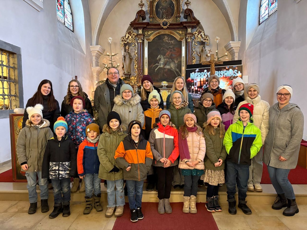 Eine Gruppe von Menschen, einschließlich Kindern, steht vor einem Altar in einer Kirche und lächelt für ein Foto. Dahinter wird ein Weihnachtsschild auf einem Bildschirm angezeigt.