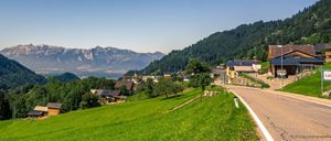 Ein ruhiges Dorf in einem alpinen Tal, umgeben von üppigen grünen Hügeln und hohen Bergen. Ein klarer Himmel und ein kleiner Fluss verstärken die friedliche Atmosphäre.