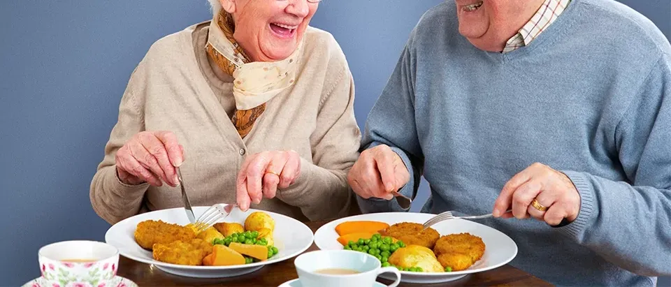 Zwei ältere Menschen lächeln und essen Essen auf weißen Tellern. Sie sitzen an einem Holztisch mit Tassen.