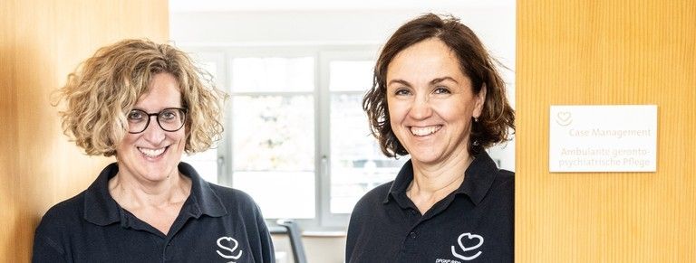 Zwei Frauen in schwarzen Hemden stehen in einem Raum mit einem Schild, das 'Case Management' und 'Ambulante geronto-psychiatrische Pflege' besagt. Hinter ihnen bietet ein Fenster einen Blick auf einen Raum mit einem Schreibtisch.