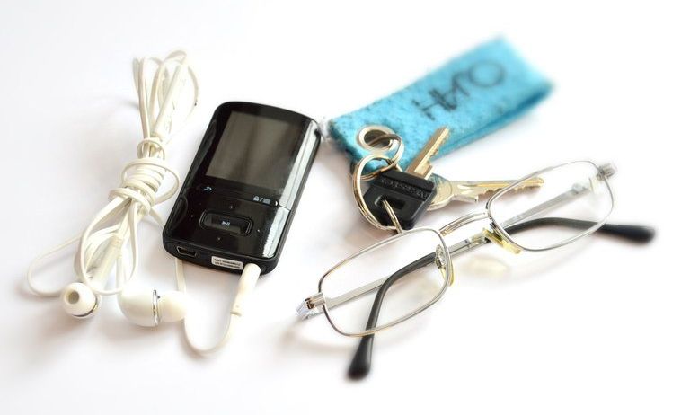 Ein schwarzer MP3-Player mit Kopfhörern, eine Brille und ein Schlüsselanhänger liegen auf einer weißen Oberfläche.