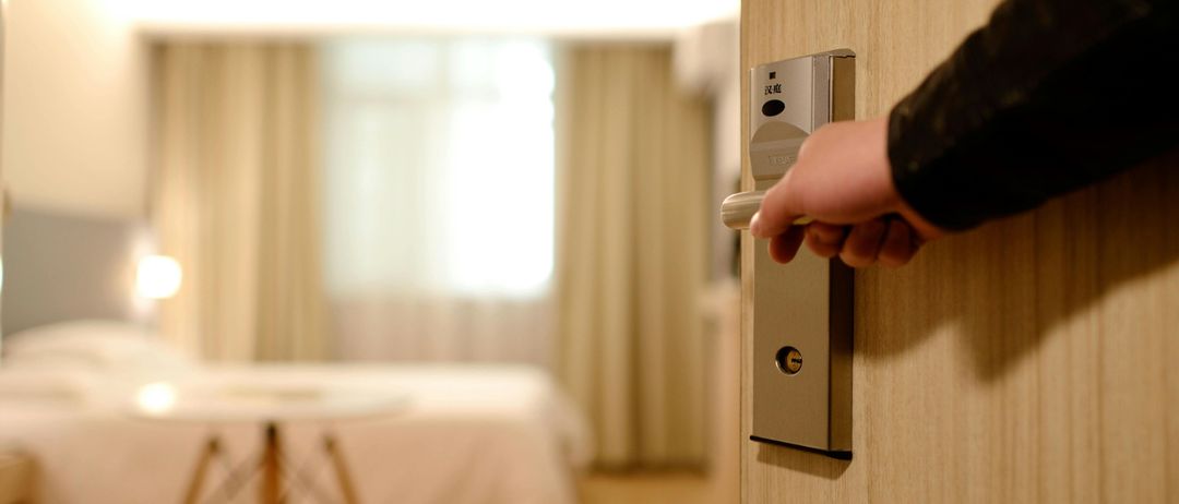 Eine Hand öffnet eine Hotelzimmertür mit einem digitalen Schloss, wobei ein Schlafzimmer und ein Tisch im Hintergrund zu sehen sind.