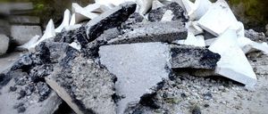 Ein Haufen Schutt, einschließlich grauer Beton und weißer Materialien, verstreut auf dem Boden mit einer moosbewachsenen Wand im Hintergrund.