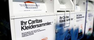 Eine Reihe weißer Container für Kleiderspenden. Jeder Container ist mit 'Ihr Caritas Kleidersammler' beschriftet. Ein Sack mit gespendeten Kleidern ist auf einem der Container sichtbar.