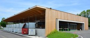 Ein modernes Gebäude mit Holzfassade und Flachdach. Mehrere weiße Container sind außen aufgestellt. Der Eingang ist weit geöffnet, und eine kleine Rasenfläche befindet sich auf der rechten Seite.