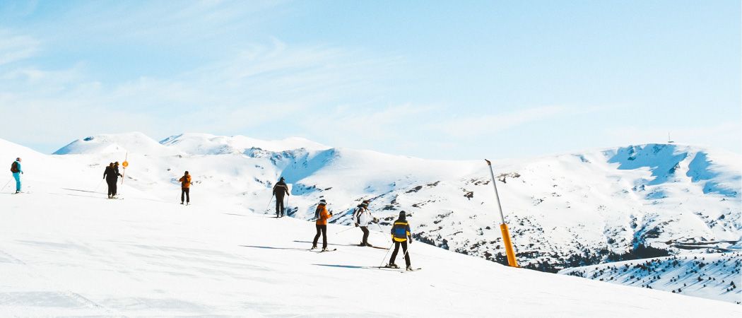Eine Gruppe von Skifahrern, in Winterkleidung gekleidet, fährt den schneibedeckten Hang unter einem blauen Himmel mit einigen Wolken hinunter.