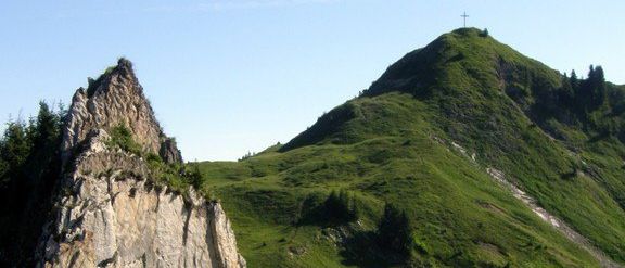 Ein felsiger Berg mit einem Kreuz oben und einem grün bewachsenen Hang mit kleinen Bäumen.