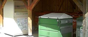 Ein Gebäude mit einem Dach aus Schindeln hat einen offenen Eingang. Drinnen steht ein weißer Spendencontainer für Kleidung. Zwei grüne Müllcontainer mit Deckeln sind im Inneren geparkt.