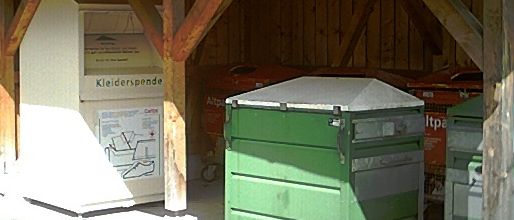 Ein Gebäude mit einem Dach aus Schindeln hat einen offenen Eingang. Drinnen steht ein weißer Spendencontainer für Kleidung. Zwei grüne Müllcontainer mit Deckeln sind im Inneren geparkt.