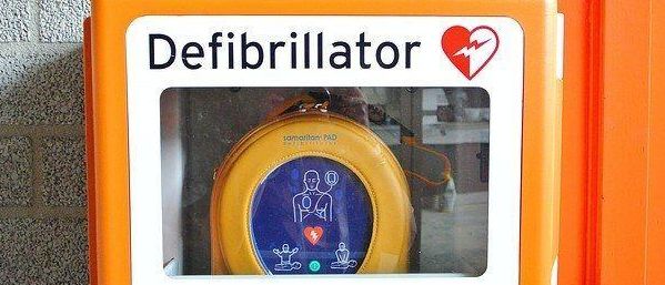 Ein orangefarbener Defibrillator-Schrank mit einem roten Herzsymbol oben drauf. Im Inneren des Schranks befindet sich ein Defibrillator.