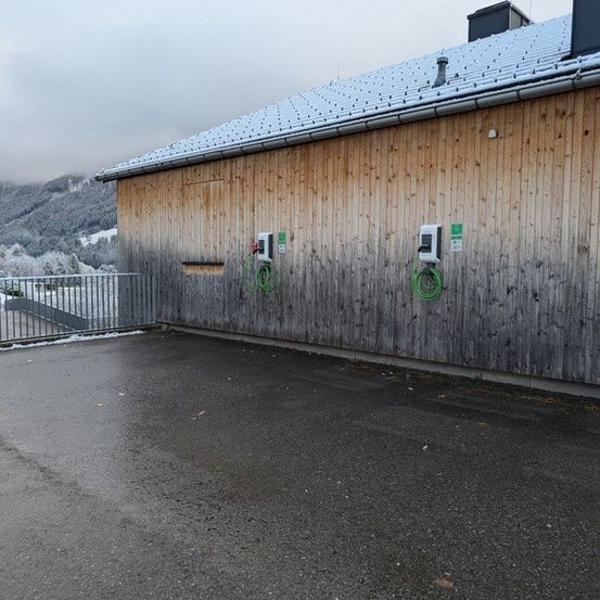 Ein verschneites Holzgebäude mit einem Metallzaun hat zwei Elektrofahrzeug-Ladestationen an seiner Wand montiert.