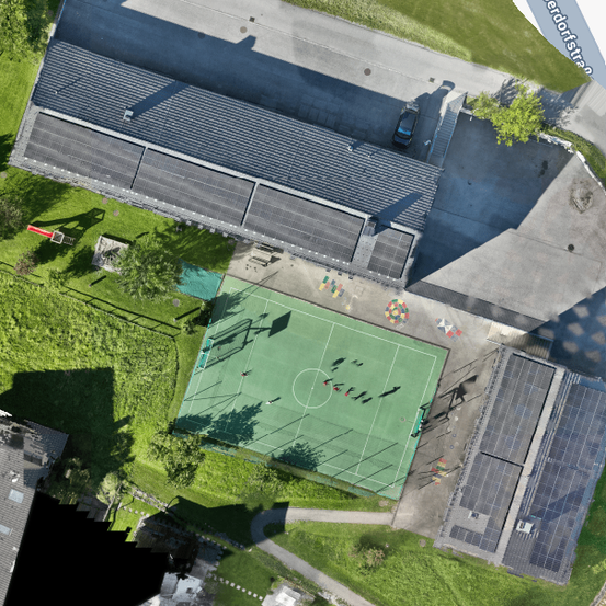 Ein Blick von oben zeigt ein Sportfeld, auf dem eine Gruppe Menschen Fußball spielt. Das Feld ist von Solarpanelen, Bäumen und Gras umgeben. Gebäude befinden sich auf beiden Seiten und ein Auto ist in der Ferne geparkt.