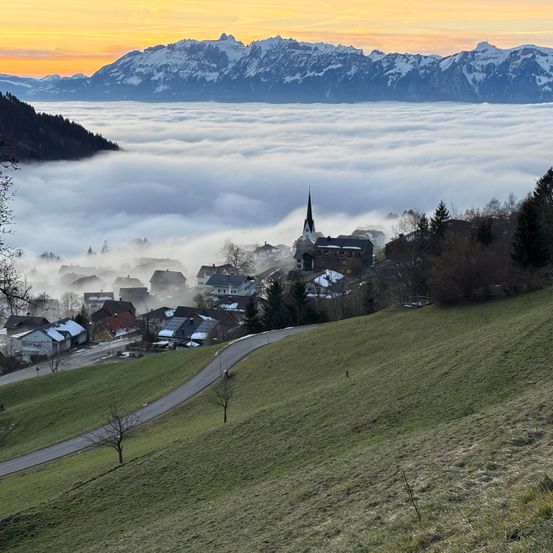 Ein in den Bergen gelegenes Dorf mit einem Turm, der sich über den Nebel erhebt. Der Himmel ist eine Mischung aus Orange und Grau.
