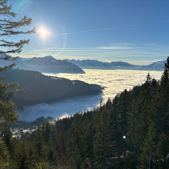 Ein Panoramablick von einem Berggipfel mit strahlend heller Sonne. Das Tal unten ist von Nebel bedeckt, und in der Ferne befinden sich schneebedeckte Berge.