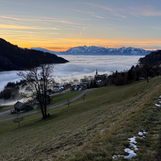 Ein nebliger Morgen in einem Tal mit einem kleinen Dorf, das zwischen den Hügeln liegt. In der Ferne sind schneebedeckte Berge sichtbar unter einem goldenen Sonnenuntergang.