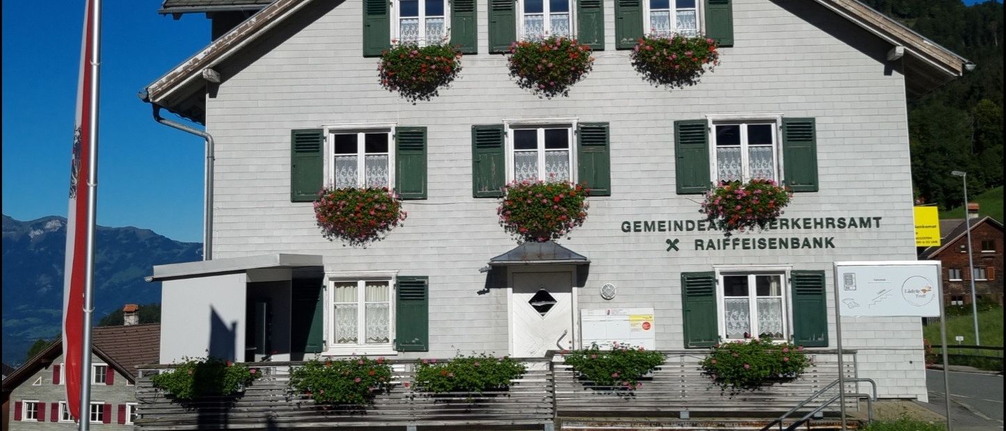 Ein zweistöckiges Gebäude mit einem dreieckigen Dach, weißen Wänden und grünen Fensterläden. Es hat eine Reihe von Blumenkästen über den Fenstern und ein Schild, das darauf hinweist, dass es sich um eine Gemeindebank handelt.