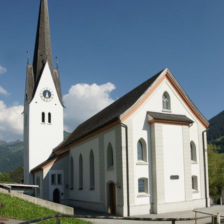 Eine weiße Kirche mit einem Turm und Uhren steht vor einem Bergpanorama. Die Kirche hat Rundbogenfenster und eine Holztür, mit einem Zaun und Gras davor.