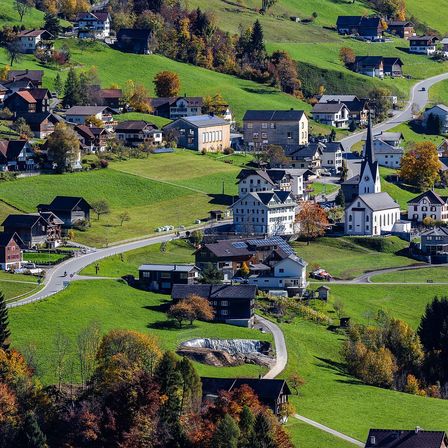 Ein landschaftlicher Luftblick auf ein kleines Dorf in einem üppigen grünen Tal, mit traditionellen Holzhäusern und einer Kirche mit einem Kirchturm.