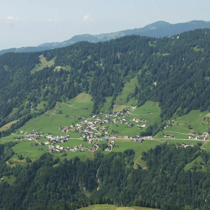 Luftaufnahme eines kleinen Dorfes in einem Tal, umgeben von dichten Wäldern und grünen Hügeln unter einem klaren Himmel.