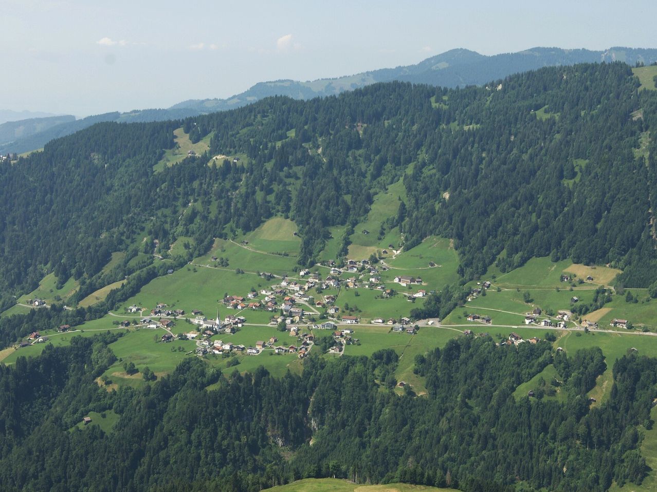 Luftaufnahme eines kleinen Dorfes in einem Tal, umgeben von dichten Wäldern und grünen Hügeln unter einem klaren Himmel.