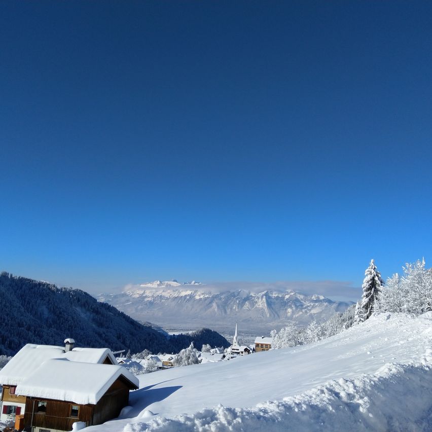 Eine verschneite Landschaft zeigt ein Haus unter einem blauen Himmel, umgeben von schneebedeckten Bäumen und Bergen. Eine Kirche ist in der Ferne zu sehen.