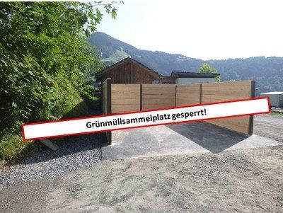 Ein roter Banner über einer Schotterauffahrt zeigt, 'Grünwiesenparkplatz gesperrt!' Die Auffahrt führt zu einem Holztor. Hinter dem Tor steht ein Haus, mit Bergen im Hintergrund.
