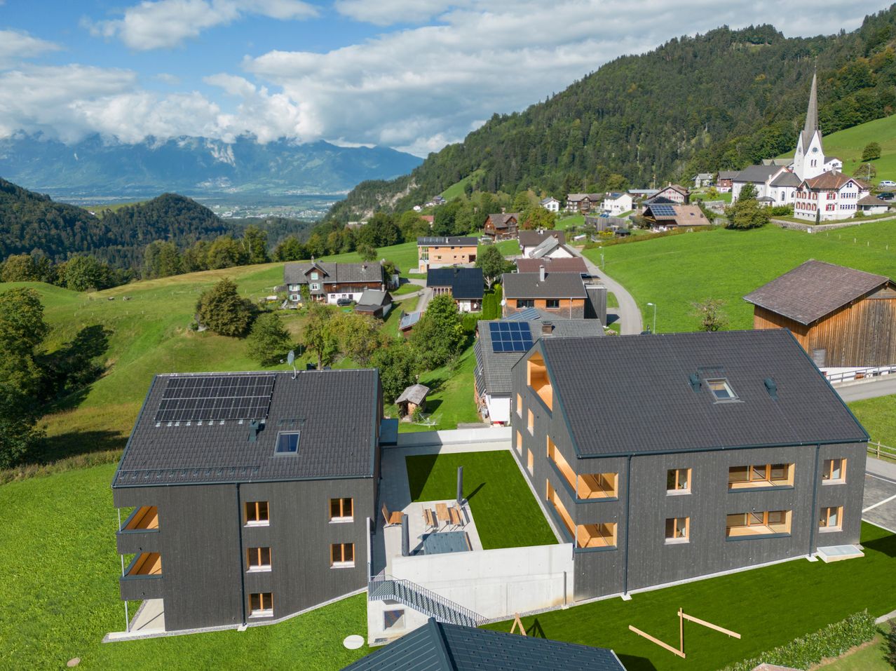Ein Luftbild einer grünen Hügellandschaft mit mehreren modernen Häusern, Solarpanels auf Dächern, umgeben von üppiger Grünfläche und Bergen in der Ferne.