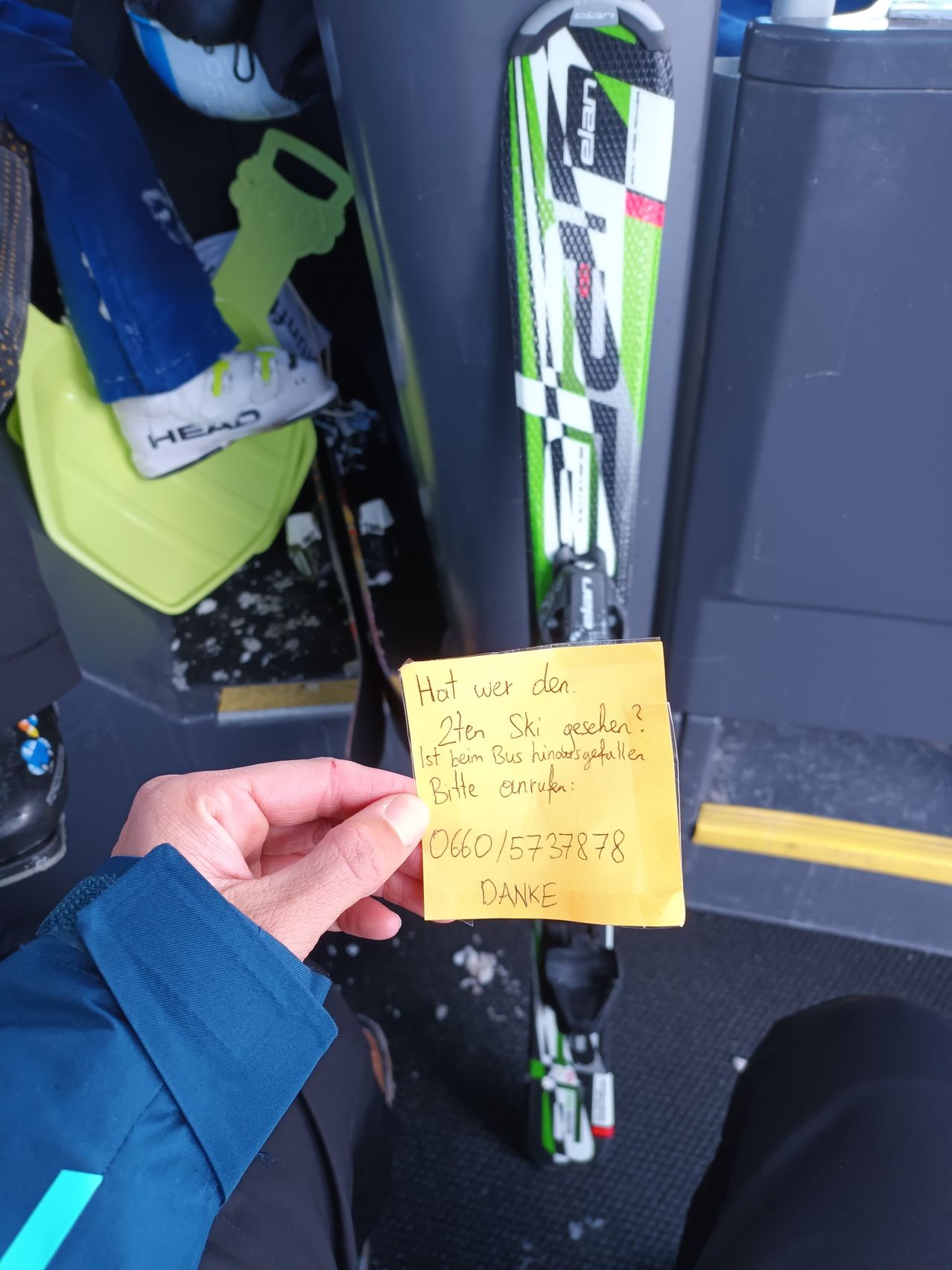 Eine Person hält eine gelbe Notiz, in der gefragt wird, ob jemand ihre Skier gesehen hat, die vom Bus gefallen sind. Die Notiz enthält eine Telefonnummer und das Wort Danke.