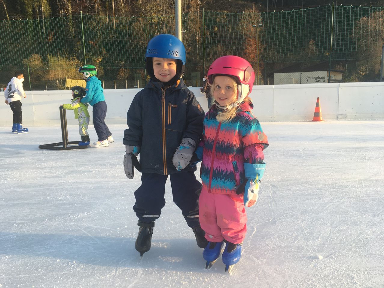 Zwei Kinder beim Eislaufen, eines mit blauem Helm, das andere mit rosa Helm. Sie stehen auf einer Eisbahn im Freien mit einer Barriere und Bäumen im Hintergrund.