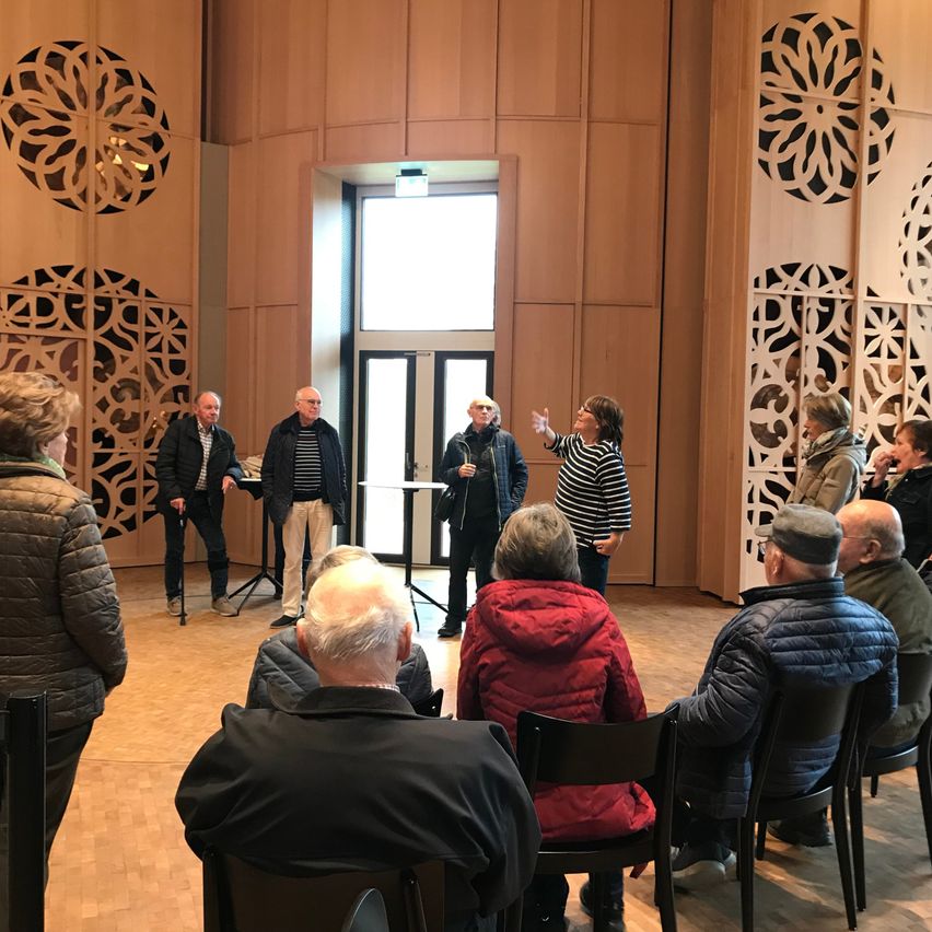 A group of people are gathered in a room, possibly for a meeting. A woman in a striped shirt stands in front of them, gesturing with her hands. The room has wooden walls and floors.