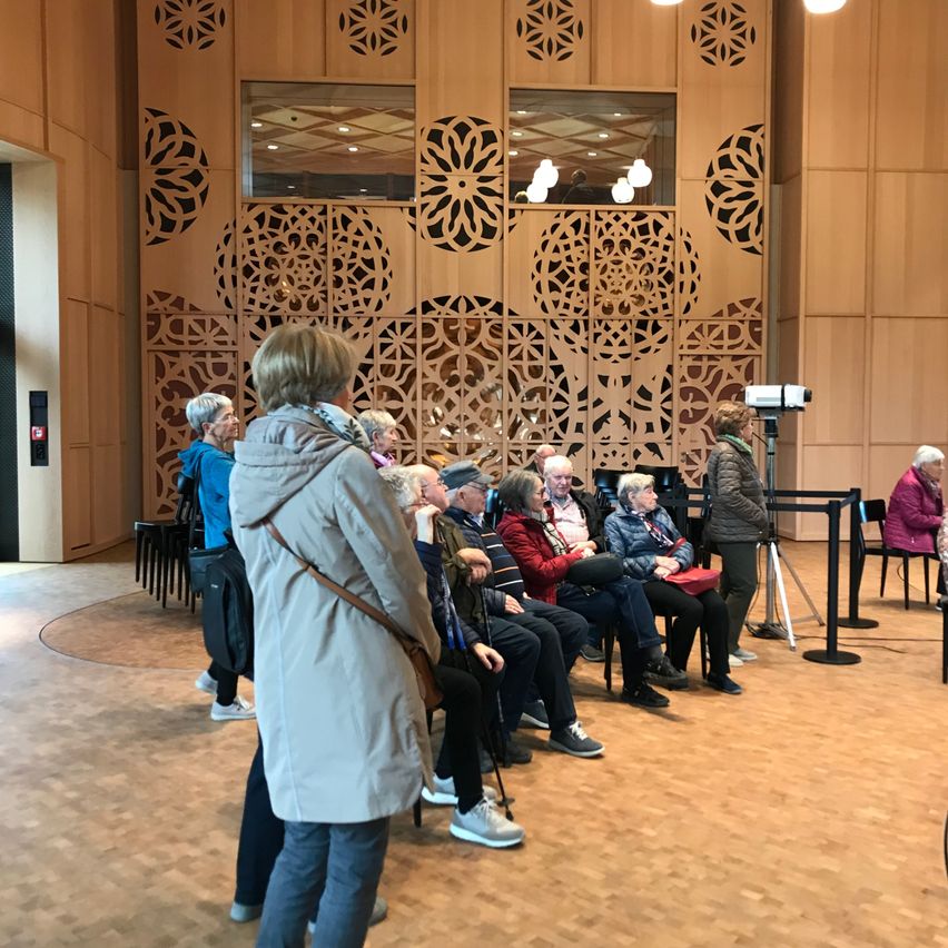 Several elderly individuals sit in chairs in a conference room with intricate wooden wall designs. One person stands near a camera, and others are engaged in conversation.