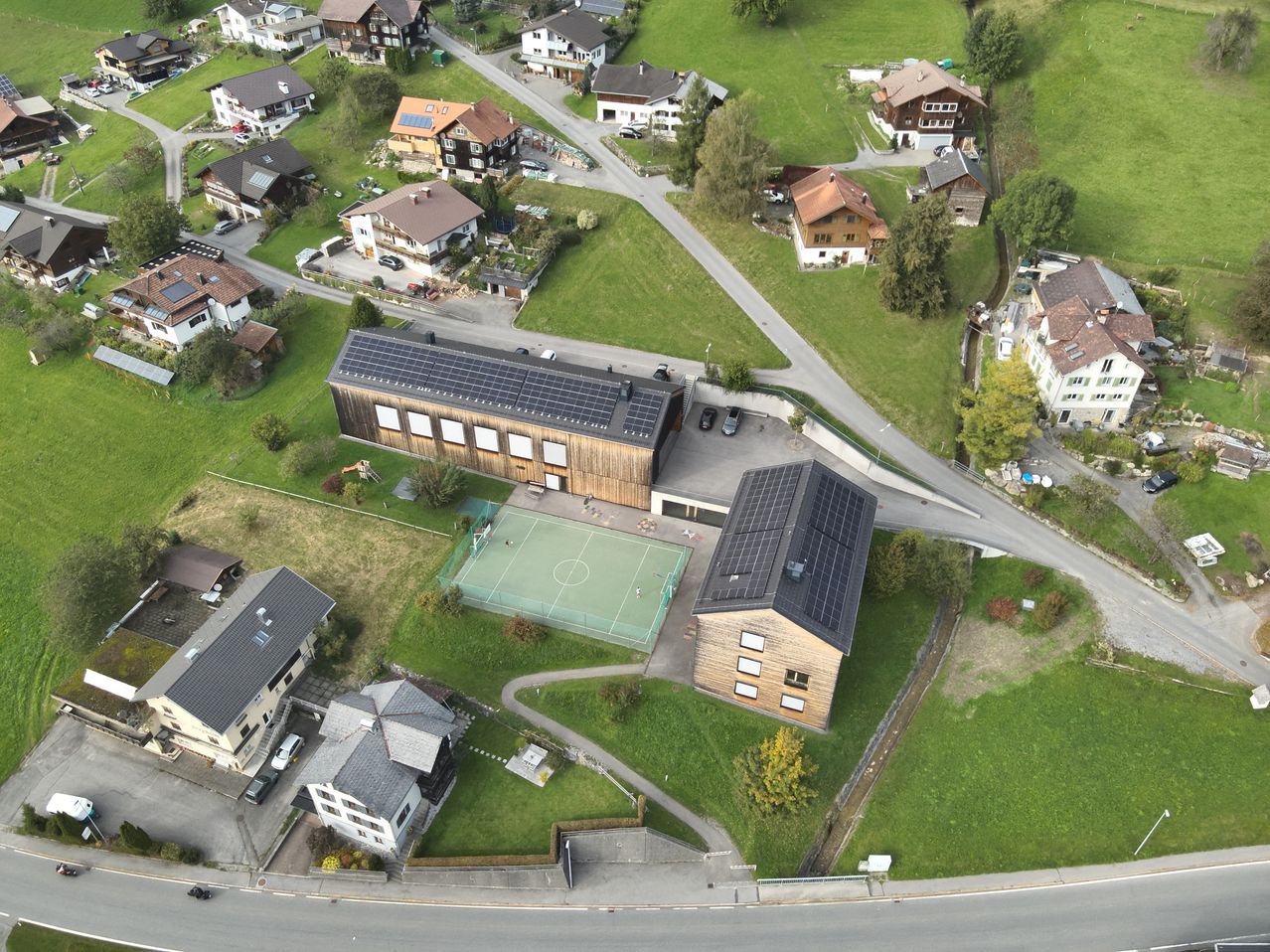 Ein Luftbild eines ländlichen Dorfes zeigt mehrere Holzhäuser, einige mit Solarpanelen auf ihren Dächern. Ein Fußballfeld mit einem grünen Netz befindet sich vor einem großen Gebäude. Das Gebiet ist von grünen Feldern und Bäumen umgeben, mit einem Weg, der zu den Häusern führt.