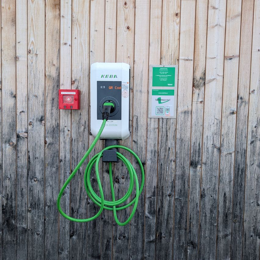 Eine Ladestation für Elektrofahrzeuge ist an einer Holzwand montiert. Sie hat einen grünen Schlauch angeschlossen. Über dem Ladegerät befindet sich ein roter Kasten mit einem QR-Code. Rechts davon befindet sich ein grünes Schild.
