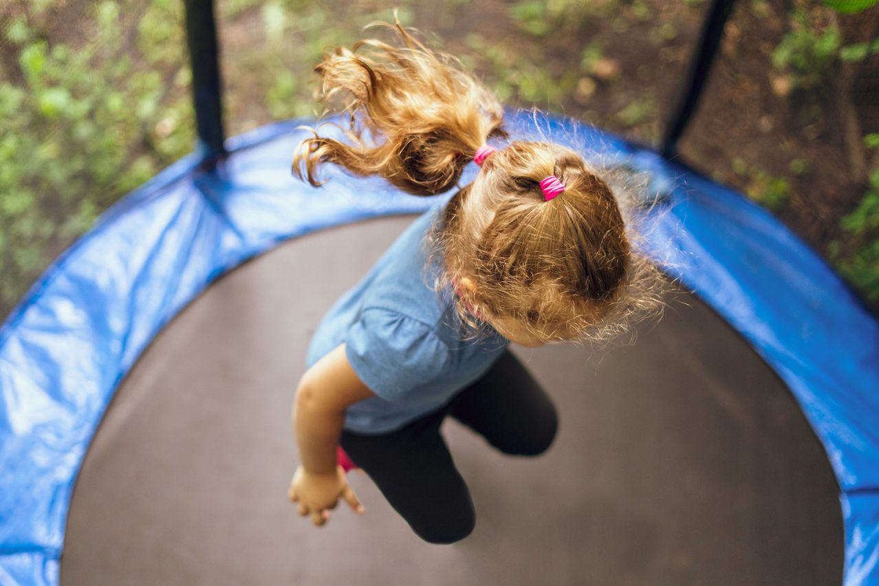 Ein junges Mädchen mit blonden Haaren springt auf einem Trampolin. Sie trägt ein blaues Shirt und schwarze Hosen.