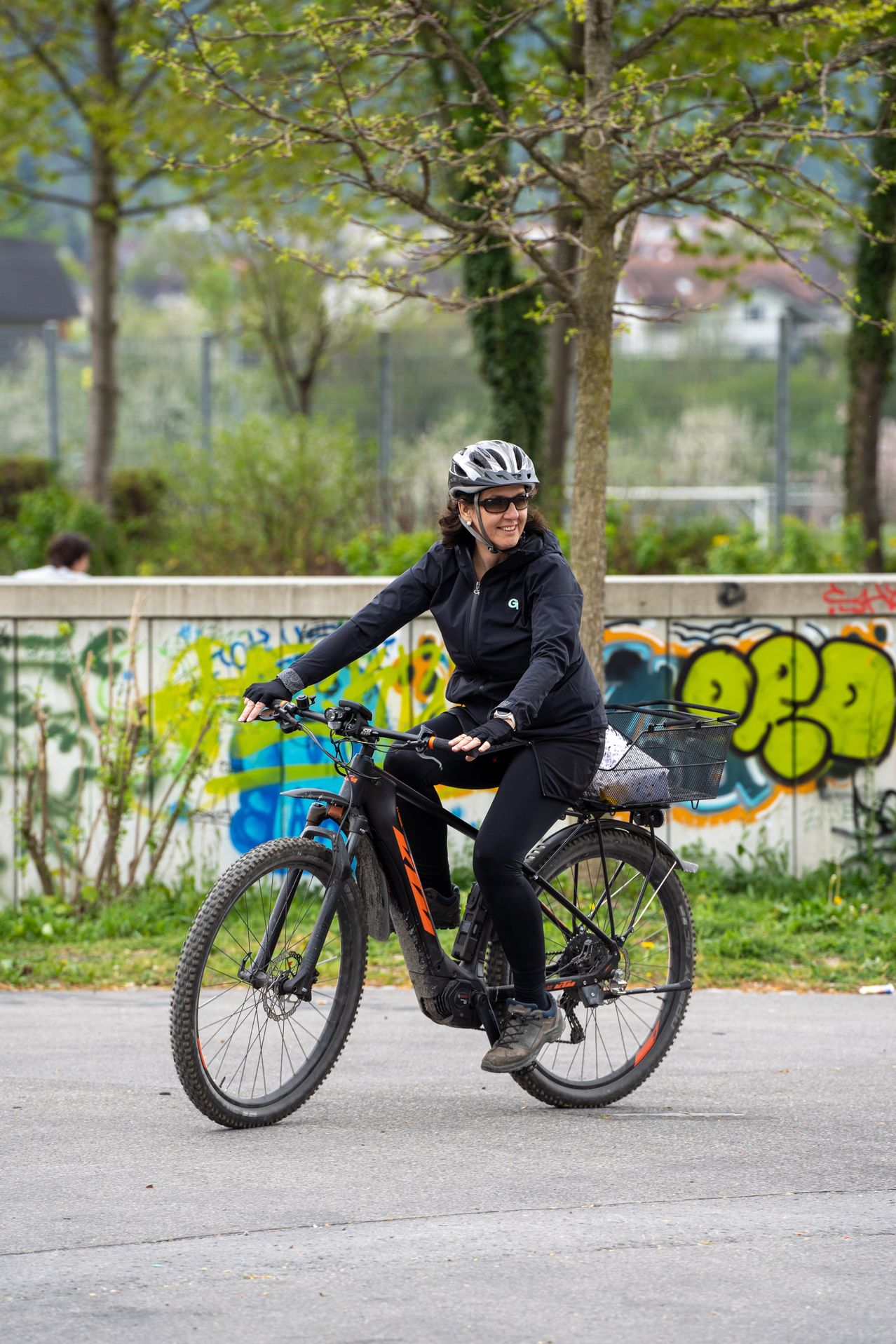 Eine Frau mit Helm fährt auf einer Straße mit Graffiti an der Wand hinter ihr ein Fahrrad. Sie trägt schwarze Kleidung und hat einen Korb am Hinterrad befestigt.