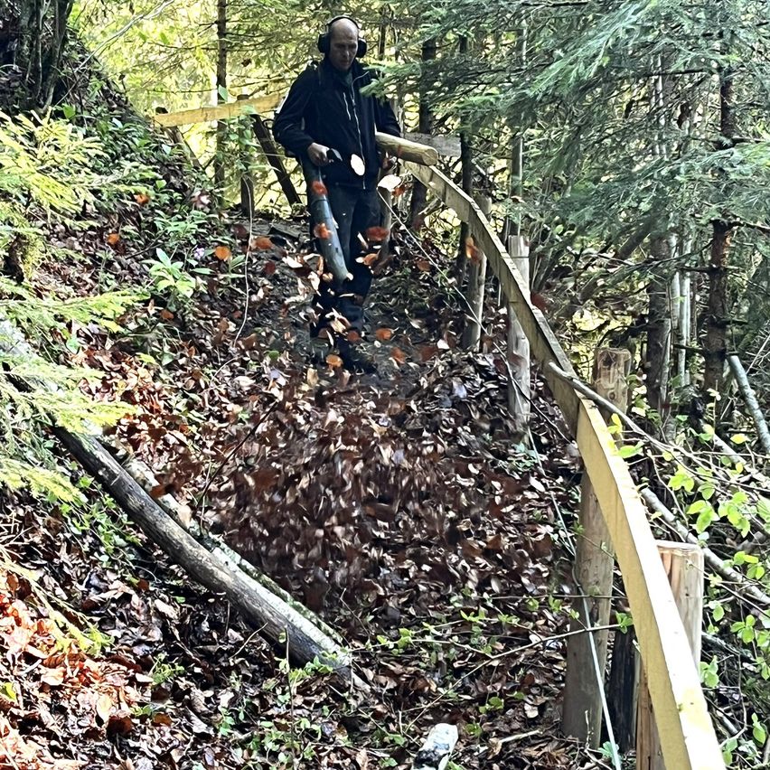 Ein Mann im Wald, mit Kopfhörern, benutzt einen Laubbläser, um Blätter von einem steilen, engen Pfad zu entfernen.