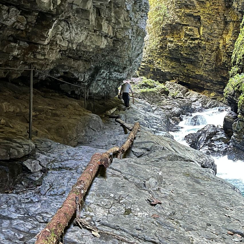 Eine Person geht auf einem schmalen Pfad an einem felsigen Abhang entlang, wobei ein Baumstamm über die Felsen gelegt wurde, um Stabilität zu gewährleisten. Der Pfad führt zu einem Wasserstrom, der durch eine Schlucht fließt.