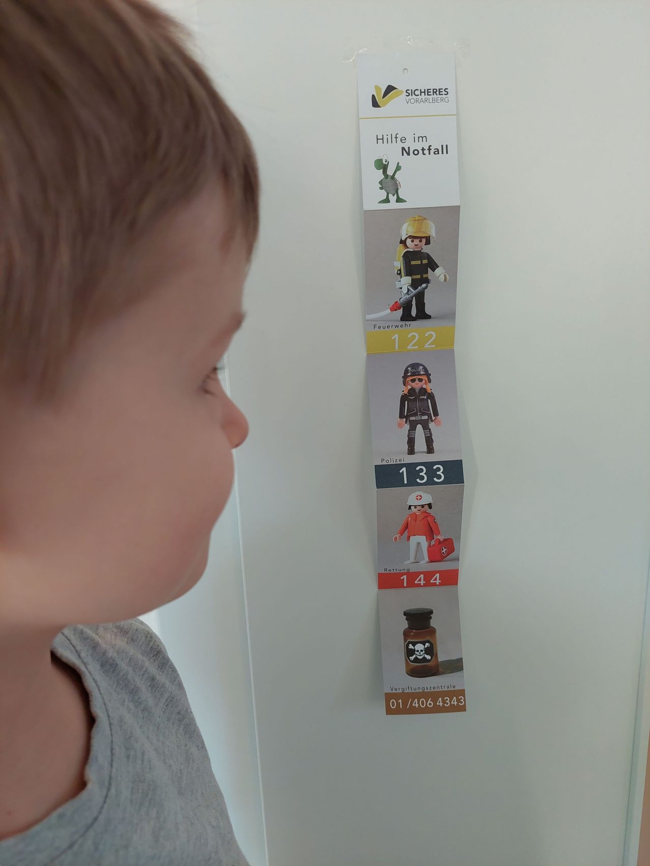 Ein junges Kind untersucht eine vertikale Liste von Notfallnummern. Die oberste Nummer ist 122, gefolgt von 133 und dann 144. Unter diesen Nummern befindet sich ein Bild einer Giftflasche mit Totenkopf und Knochenkreuz. Der Hintergrund ist weiß.