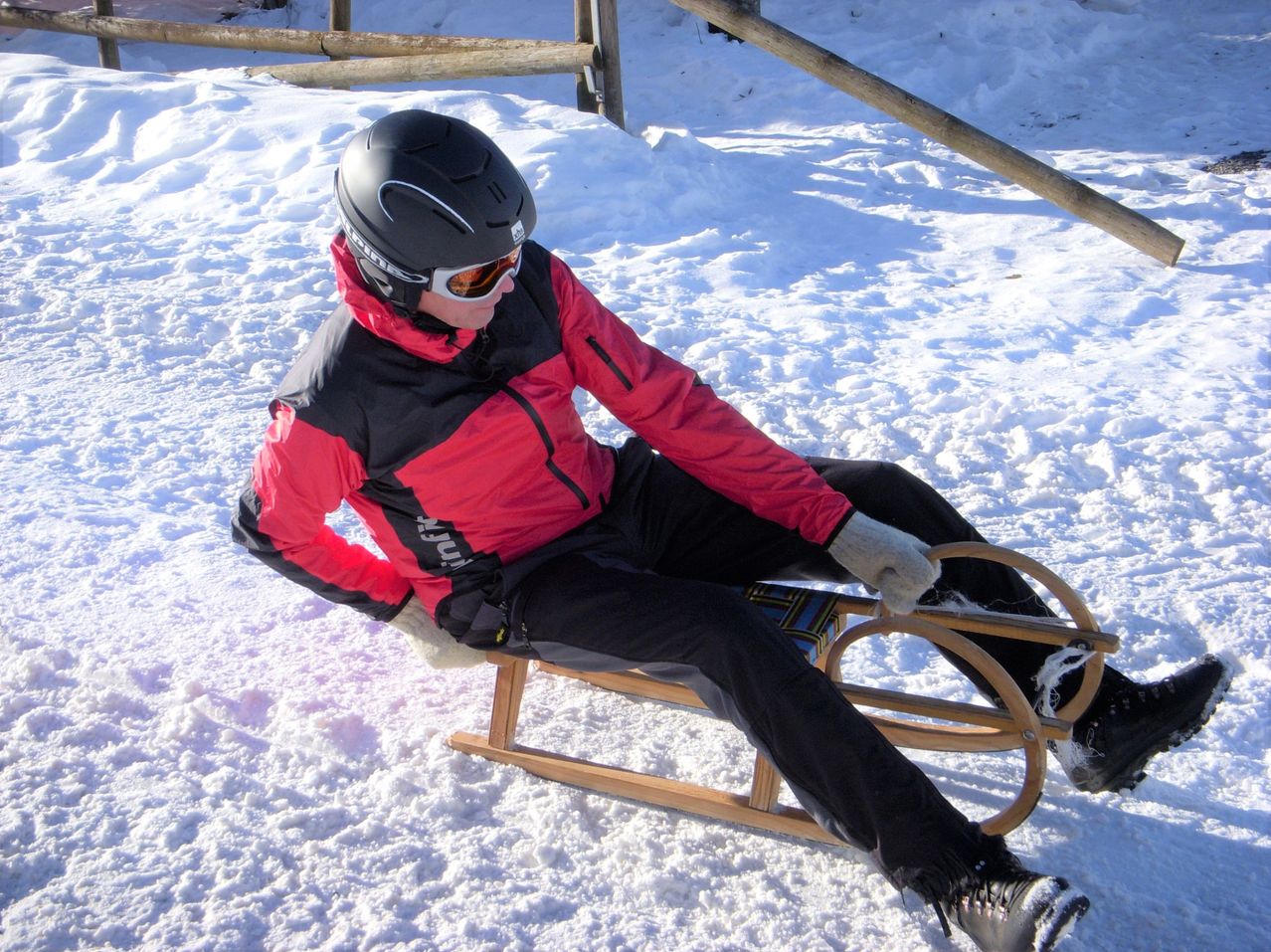 Eine Person sitzt auf einem hölzernen Schlitten im Schnee, trägt eine rot-schwarze Jacke, schwarze Hose und einen schwarzen Helm. Der Schlitten hat zwei Griffe auf jeder Seite.