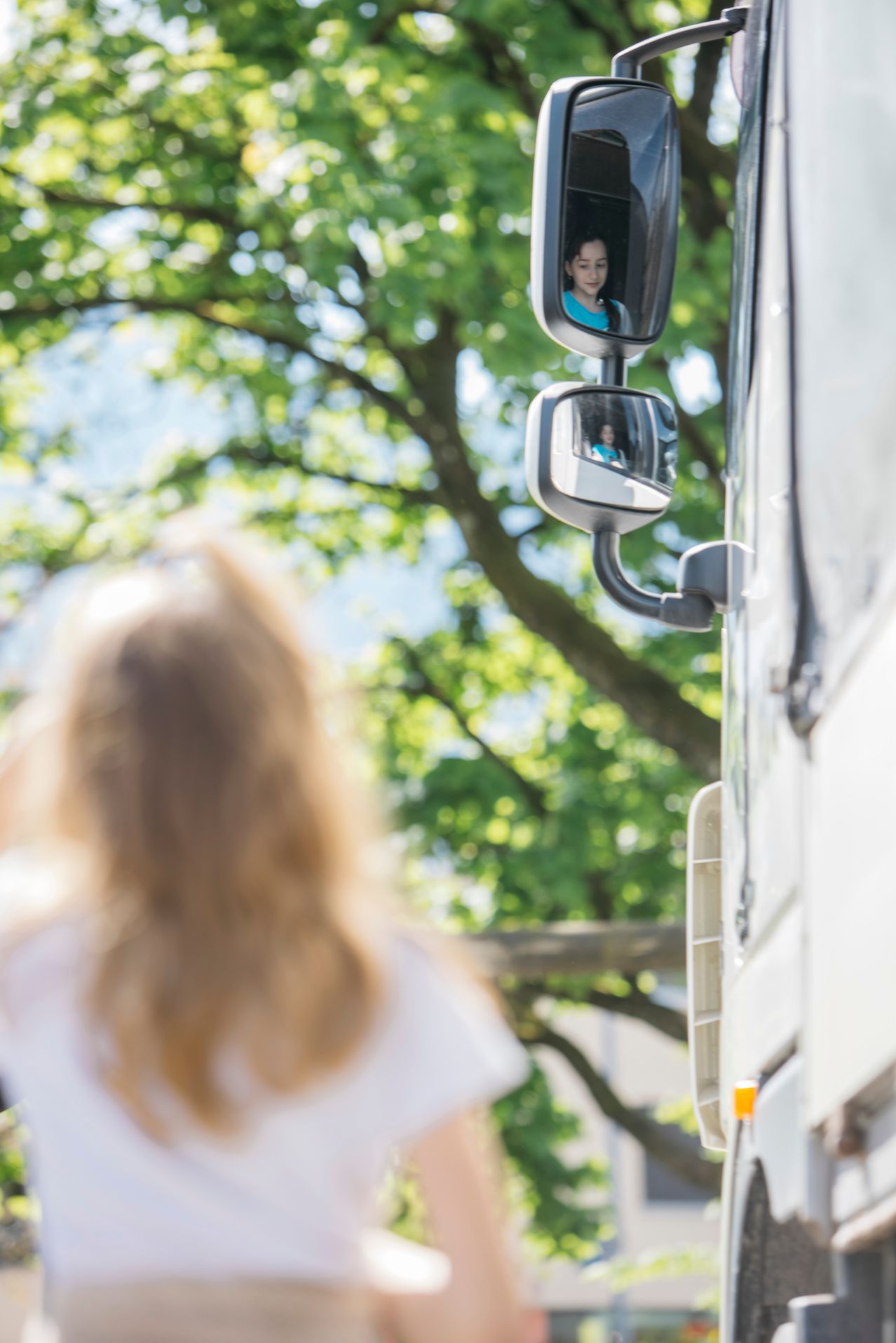 Der Rückspiegel eines Fahrzeugs zeigt die Reflexion einer Frau, während eine weitere Frau mit langen Haaren im Vordergrund steht.