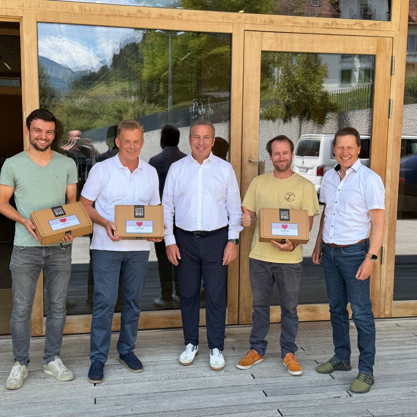 Fünf Männer stehen vor einer Holztür, lächelnd und Boxen mit einem Herzdesign in den Händen. Sie sind in Freizeitkleidung gekleidet. Hinter ihnen spiegelt eine Glastür eine Landschaft mit Bergen und Bäumen.