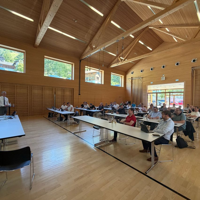 Ein Konferenzraum mit Holzwänden und -decke, gefüllt mit Menschen, die an Tischen sitzen. Ein Mann steht vorne, möglicherweise leitet er die Besprechung.