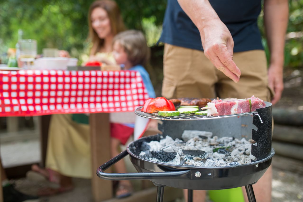 Ein Mann grillt Fleisch und Gemüse auf einem Grill, während eine Frau und ein Kind am Tisch sitzen.