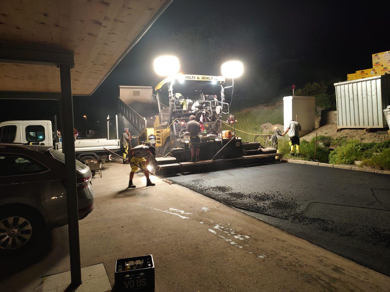 Nachtarbeiten mit einer Hilti & Jehle-Maschine. Arbeiter verteilen Asphalt auf einer Straße. Lichter und Fahrzeuge sind sichtbar.