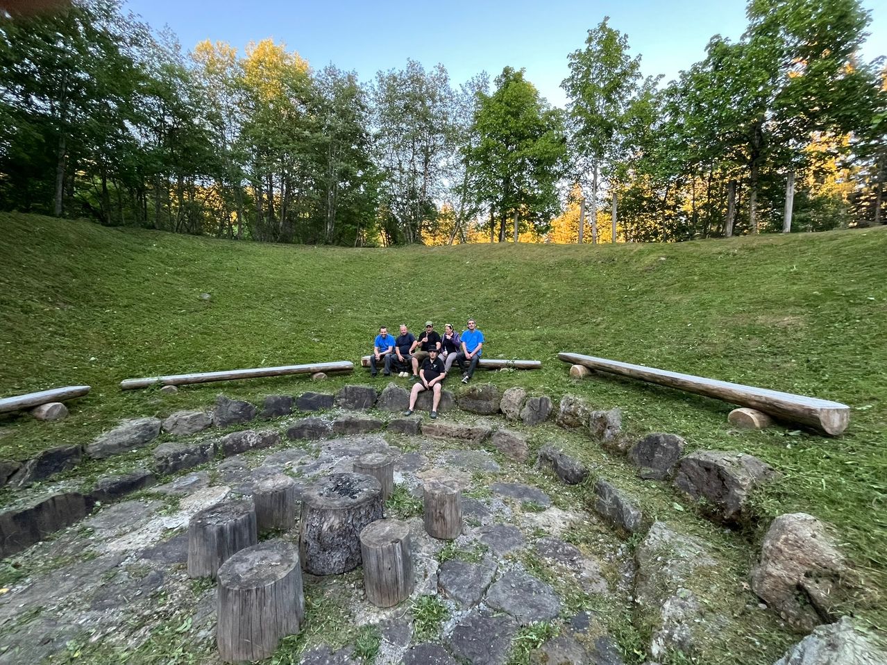 Eine Gruppe von Menschen sitzt auf Baumstümpfen in einem von Bäumen umgebenen Grasgebiet und posiert für ein Foto.