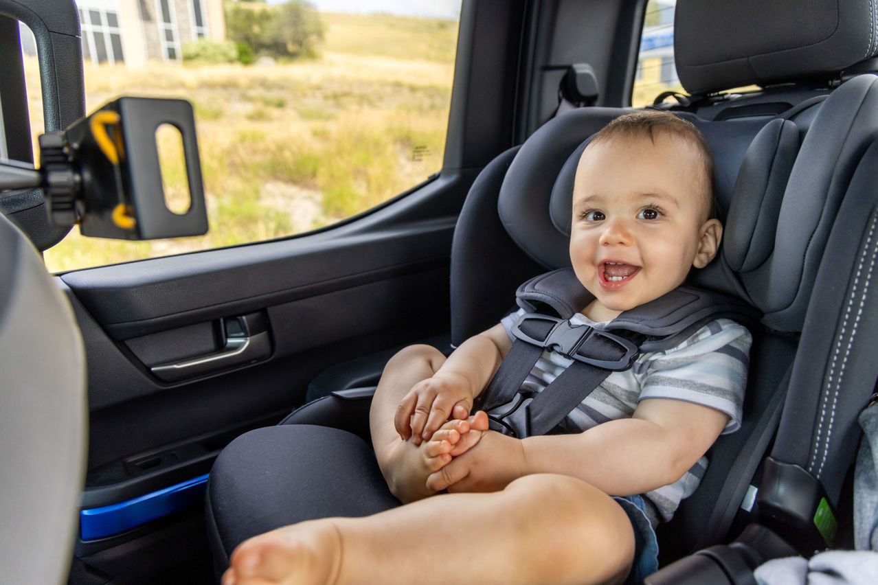 Ein Baby im Autositz lächelt mit gespreizten Beinen in einem Fahrzeug mit Blick auf ein Grasfeld.